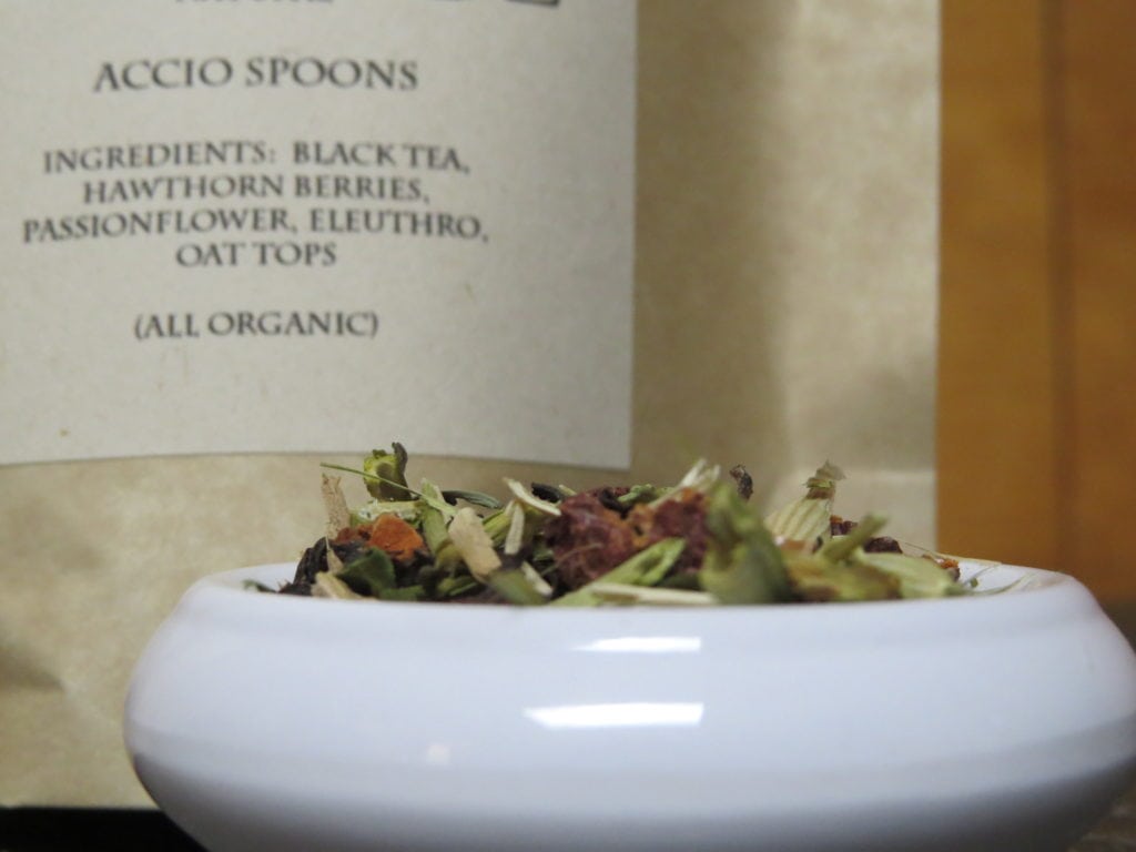 A closeup image of a small white bowl full of a blend of passionflower, eleuthro, oat tops, hawthorn berries, and black tea. Behind it is the slightly blurry label that indicates what the tea is. It calls it "Accio Spoons," lists the ingredients, and indicates that they are all organic.
