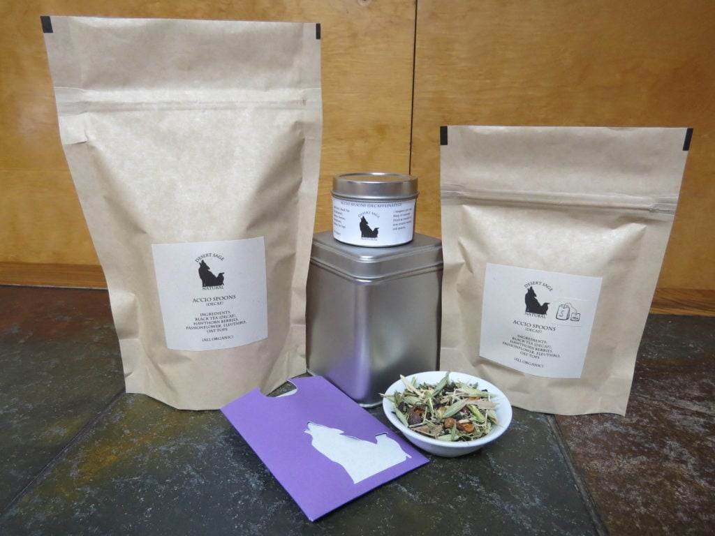 A small white bowl of tea full of herbs. Around it are arrayed a variety of tea containment objects - tins and bags, and a small rectangular packet that could hold a single teabag. They are labeled "Accio Spoons (Decaffeinated), Ingredients:Black Tea, Hawthorn Berries, Passionflower, Eleuthro, Oat Tops (All Organic)"
