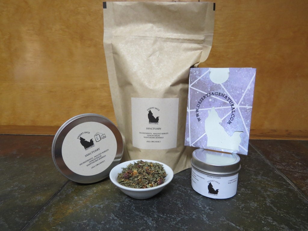 A large bag of tea, with a circular sample tins to either side, the smaller of the two has a purple packet with the Desert Sage Natural Logo as a cutout balanced on top. In front of the array is a small white bowl full of a blend of hawthorn berries, lemon balm, and barley. The labels read: "Sanctuary, Ingredients: Malted Barley, Lemon Balm, Hawthorn Berries, All Organic."