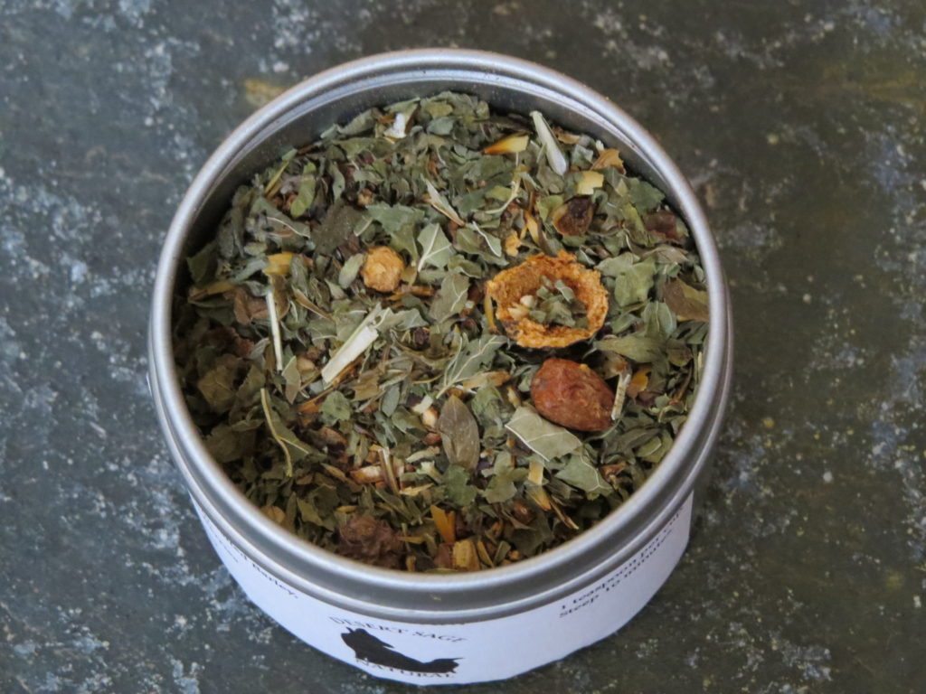 A small circular metal tin, filled with a blend of Lemon Balm, Hawthorn Berries, and Malted Barley. The shot is from above the tin, so you can clearly see the herbs, as well as the textured green marble table beneath the tea.