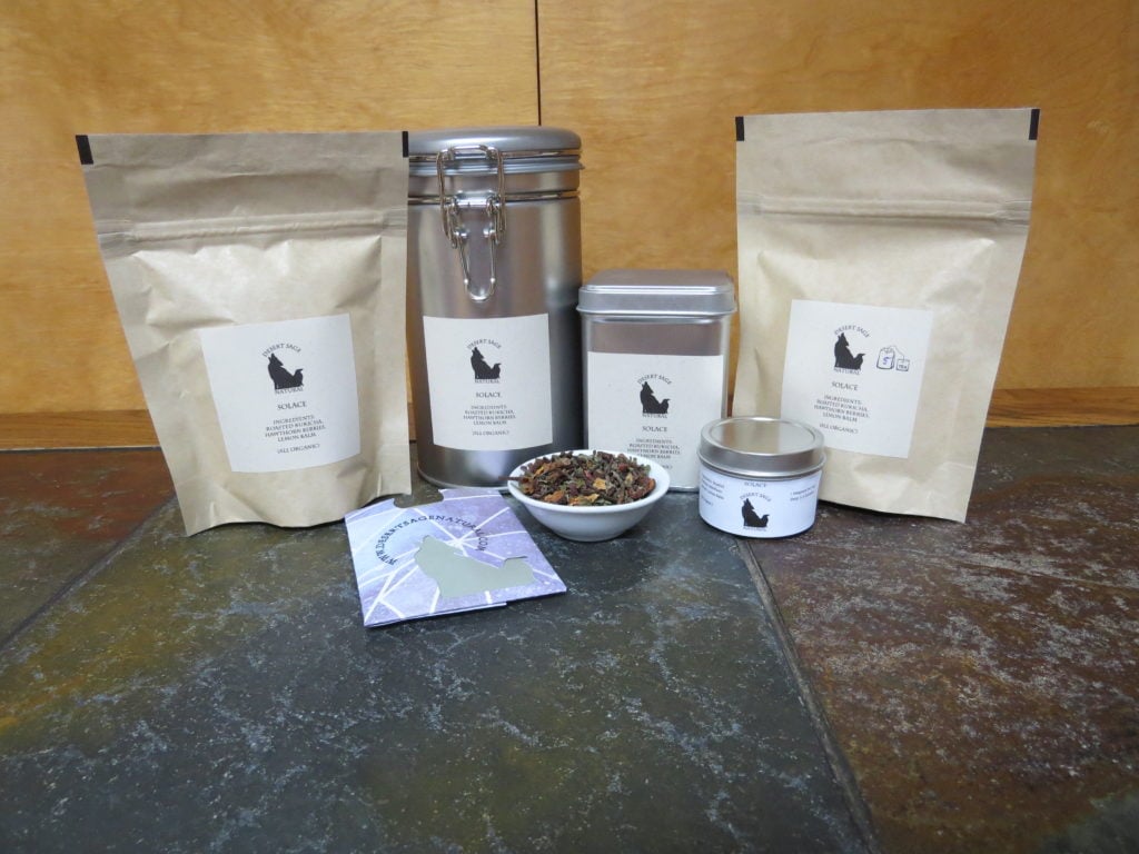 Two bags, a large latch-top tin, a standard tea tin, a small round tin, and a rectangular packet of a size to hold a teabag arrayed around a small white bowl full of a blend of roasted kukicha, hawthorn berries, and lemon balm. The labels read Solace - and list what is in the bowl as ingredients, specifying that they are organic.