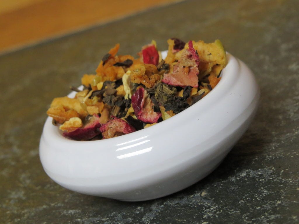 Closein shot of a small white bowl on a textured stone table. It is taken at an angle, so the bowl fills the frame diagonally. It is full of a blend of black tea, cranberries, apple, and spices.