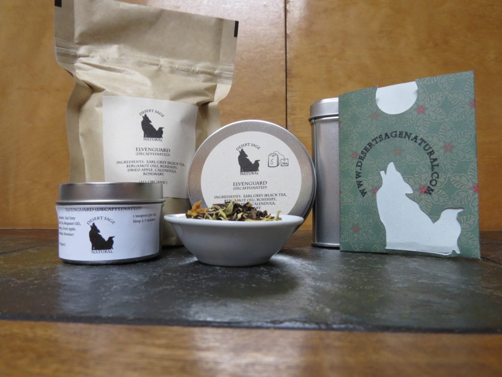 A small white bowl set on a textured green marble tabletop, the light catching the tea tin just behind so the bowl has a halo of light. The bowl is full of a mix of flowers, fruit, and tea. To the left of the bowl is a small tea tin, to the right a rectangular packet with the Desert Sage Natural Raven and Coyote logo as a cutout and www.desertsagenatural.com ringing the coyote's head. A bit further back behind is a large bag of tea. The labels read: "Elvenguard (Decaffeinated), Ingredients: Earl Grey, Rosehips, Calendula, Dried Apple, Rosemary, (All Organic)"