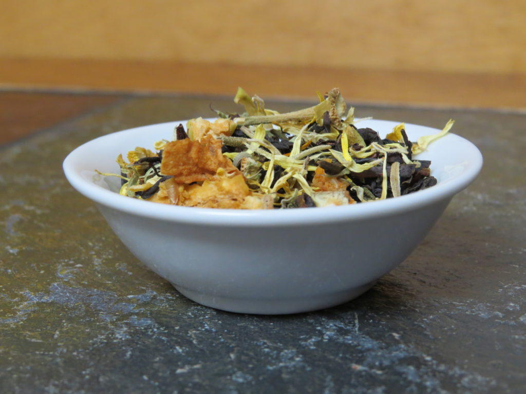 A small white bowl sitting on a green textured marble table. The bowl is full of a blend of black tea, rosehips, calendula, dried apple, and rosemary.