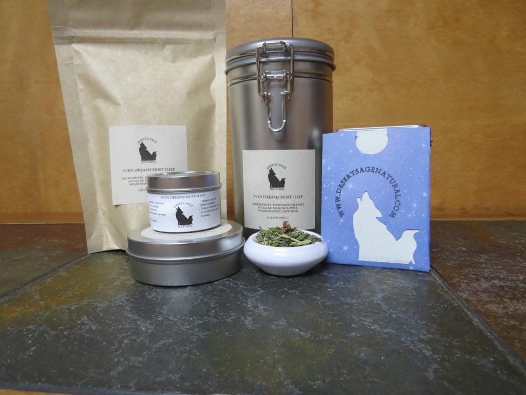 A small white bowl on a textured marble table. The bowl is full of a blend of lavender, hawthorn berries, and green herbs. To the left side are two small tins, stacked, one thinner and taller than the other. To the right is a rectangular packet with the Desert Sage Natural logo and website, of a size where it could hold a teabag. Behind the bowl is a large bag and a large tin. The labels on things read: "Even Dreams Must Sleep, Ingredients: Hawthorn Berries, Scullcap, Passionflower, Meadowsweet, Lavender, (All Organic)"