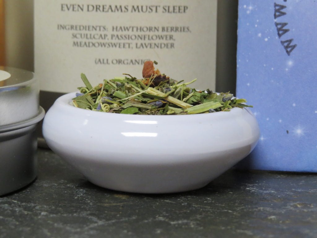 A close-up shot of a small white bowl of tea, full of a blend of lavender, hawthorn berries, and green herbs. Behind it you can just make out the words on a slightly blurred label "Even Dreams Must Sleep, Ingredients: Hawthorn berries, Scullcap, Passionflower, Meadowsweet, Lavender, (All Organic)."