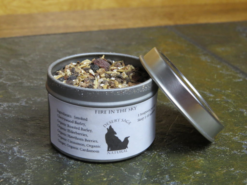 A small metal tea tin, set on a textured green stone table. The tin is open, the lid propped up at an angle on one side of it, and there is a slight dusting of barley and spice around the rim of the tin. The label reads "Fire in the Sky, Ingredients: Smoked Cherrywood Barley, Organic Roasted Barley, Organic Elderberries, Organic Hawthorn berries, Organic Cinnamon, Organic Ginger, Organic Cardamom"