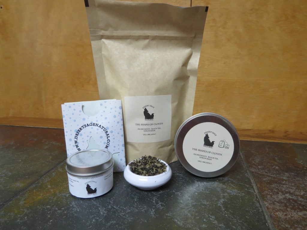 A small white bowl of tea filled with a blend of lemon balm and black tea. To either side are tea tins, behind it a large bag and a small rectangular packet that looks as if it could hold a teabag. The labels read: "The Shapes of Clouds, Ingredients: Black Tea, Lemon Balm, (All Organic).