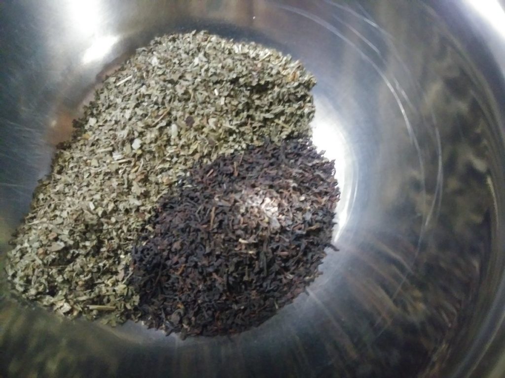 Closeup image of the inside of a metal bowl, you can only see hints of one edge - with lemon balm and black tea piled in a bowl next to each other. The herbs reflect in the side of the bowl, too.