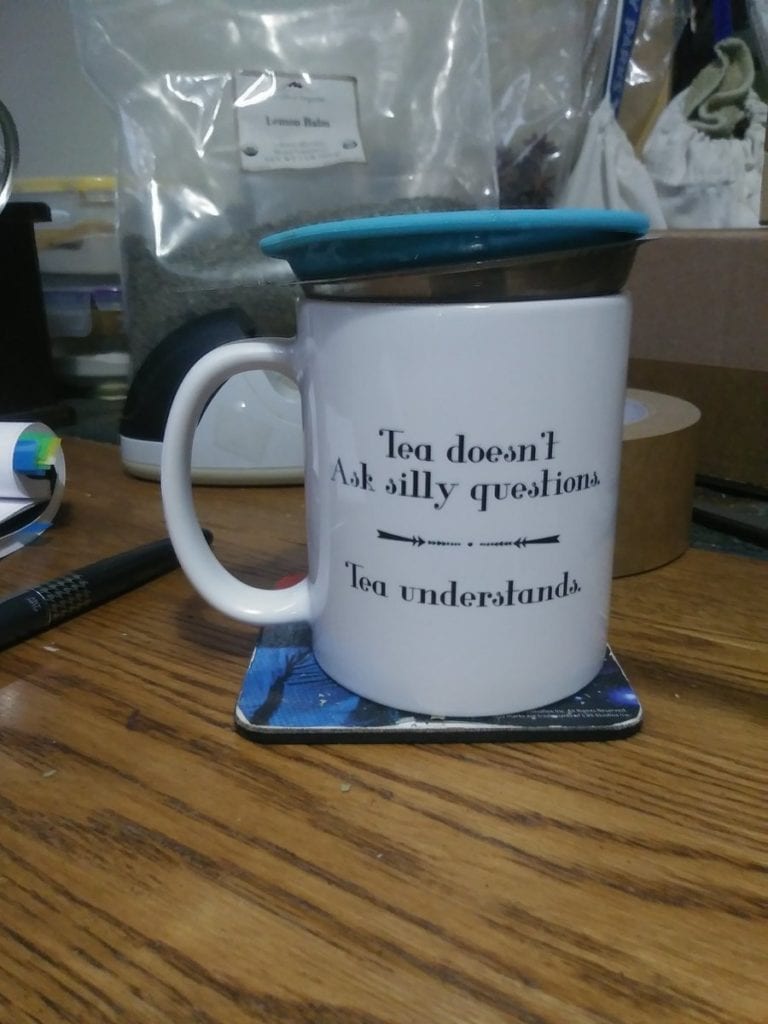 A white tea mug with the words: "Tea doesn't ask silly questions, tea understands, In it is a tea strainer with a blue lid. There is a grey pilot fountain pen to the left, a large bag of lemon balm behind, and there is a part of a roll of packing tape behind the mug, too.