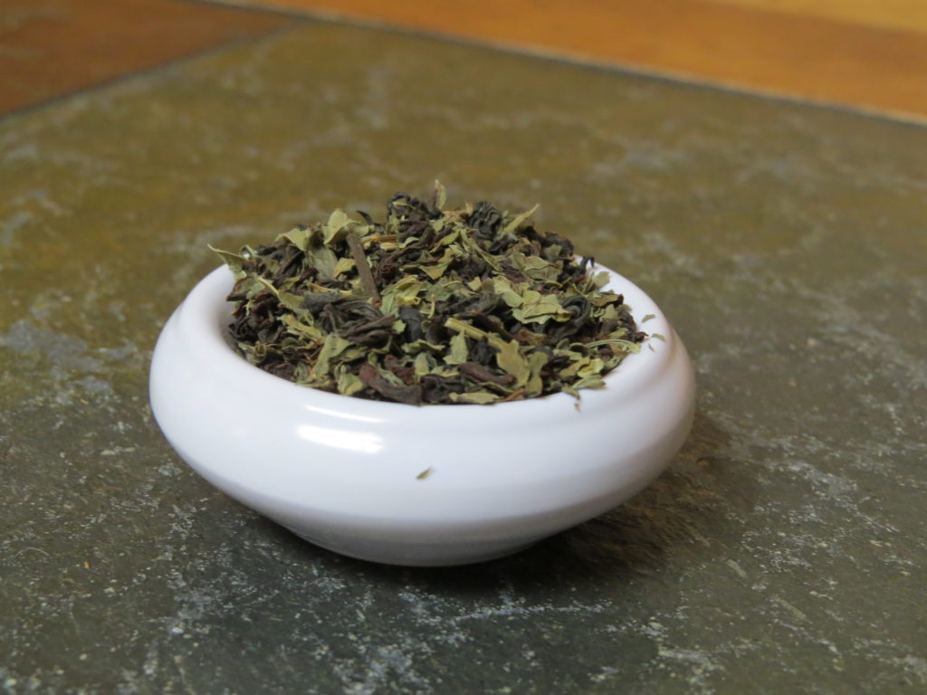 A small white bowl almost overfilled with a blend of black tea and lemon balm. It is set on a marble and wood table, the image pulled in close on the tea.