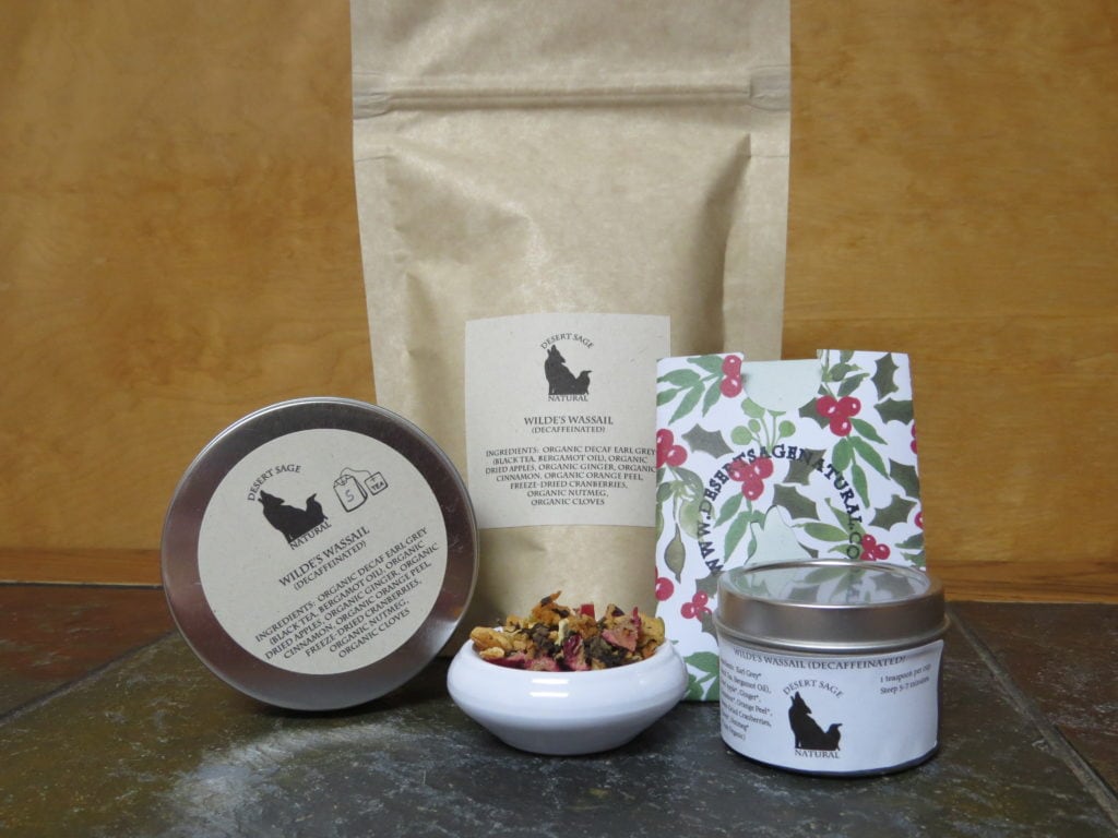 A small white bowl on a textured stone table, the background behind being a mid-range small grained wood. Behind the bowl is a bag, to the left is a circular tin up on edge, to the right is a rectangular packet with the Desert Sage Natural logo of a size to hold a teabag and a smaller circular tin. The labels read " Wilde's Wassail (Decaffeinated), Ingredients: Earl Grey, Dried Apple, Ginger, Cinnamon, Orange Peel, Freeze-Dried Cranberries, Nutmeg, Cloves" It also indicates that everything except the Cranberries are organic.