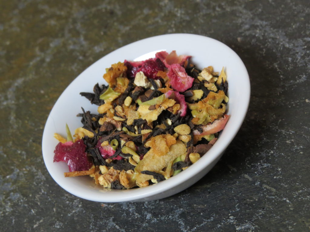 Close in shot of a small white bowl on a green textured stone table. The bowl is filled with a blend of black tea, dried apple, dried cranberries, and spices.
