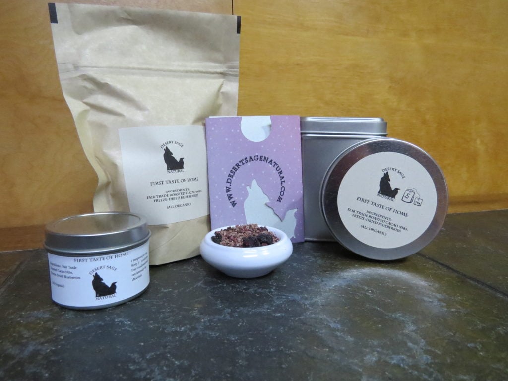 A bag of tea, two circular tea tins, a taller square tin, and a teabag packet arrayed around a small white bowl full of a combination of cacao nibs and blueberries. The labels read "First Taste of Home" and list those two ingredients, as well as stating that they are both organic.