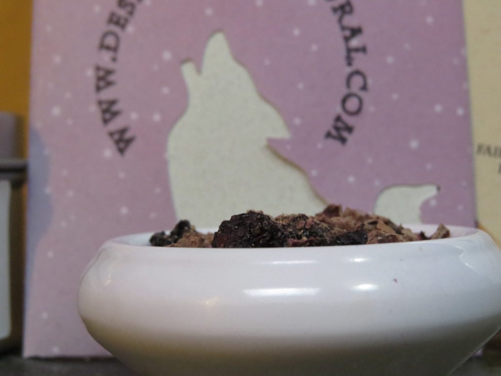 Closeup image of a small white bowl filled with ground cacao nibs and freeze dried blueberries. Behind it there is a light pinkish purple packet, with the raven and coyote logo as a cutout, and part of the web address for Desert Sage Natural.